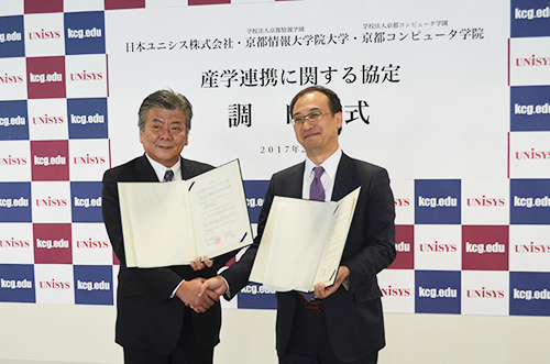 KCGI President Wataru Hasegawa (left) and Akihiro Haneda, General Manager of Nihon Unisys Research Institute, shake hands after signing an agreement on academic and research cooperation and industry-academia collaboration at KCGI Kyoto Ekimae Satellite on February 15, 2017.